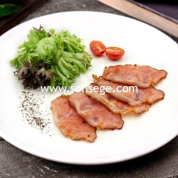 5 slices of golden brown cooked back bacon on a white round plate with lettuce and cherry tomatoes as garnish.
